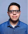 A young man in a blue button-up shirt with black glassing smiling in front of blue background