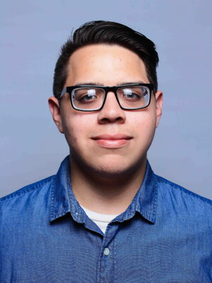 A young man in a blue button-up shirt with black glassing smiling in front of blue background